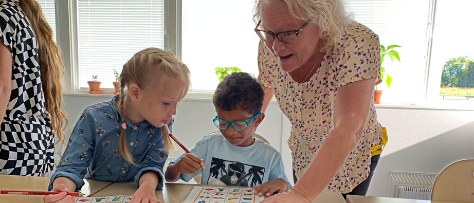 Två barn ritar på en bingobricka med bilder med hjälp av sin stödlärare
