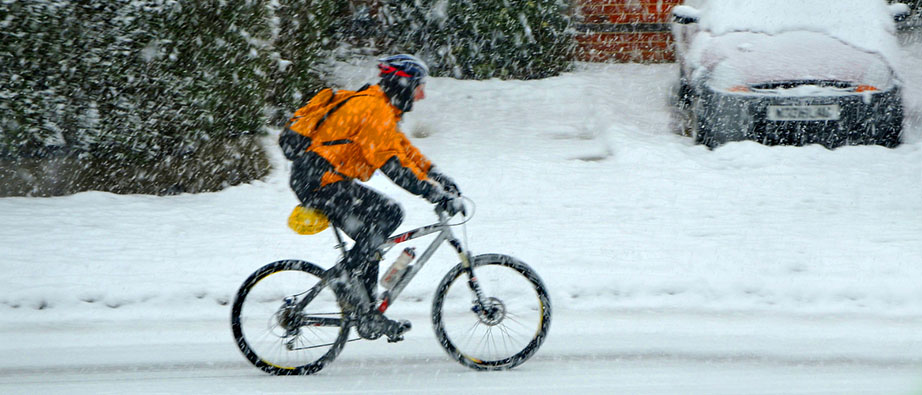 Vintercyklist på mountainbike i villaområde. Det snöar och i bakgrunden syns en översnöad bil. Foto vintercyklingens dag
