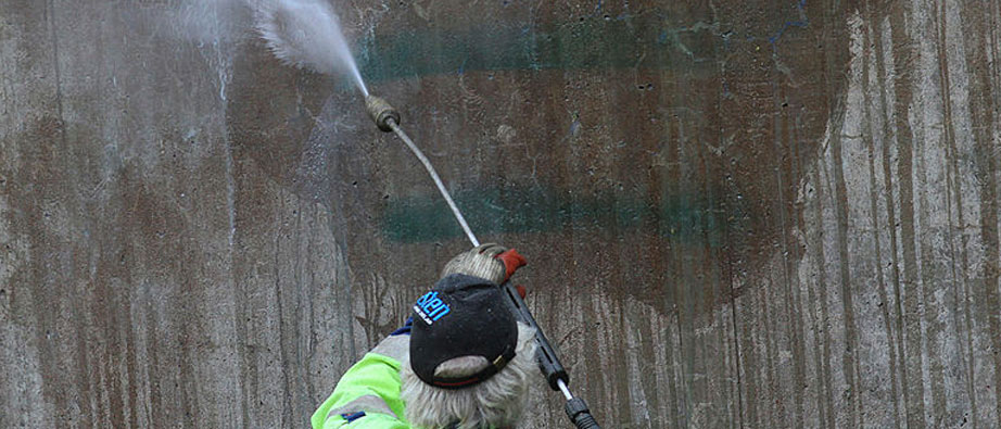 En man i arbetskläder står och spolar vatten mot en vägg för att få bort klotter.