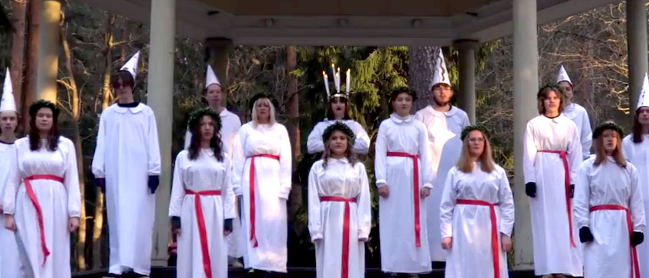 Lucia med tärnor står i vitklädda dräkter och sjunger