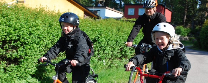 Ebba och Love cyklar glada iförda hjälmar till skolan. I Bakgrunden cyklar pappa Mats. 