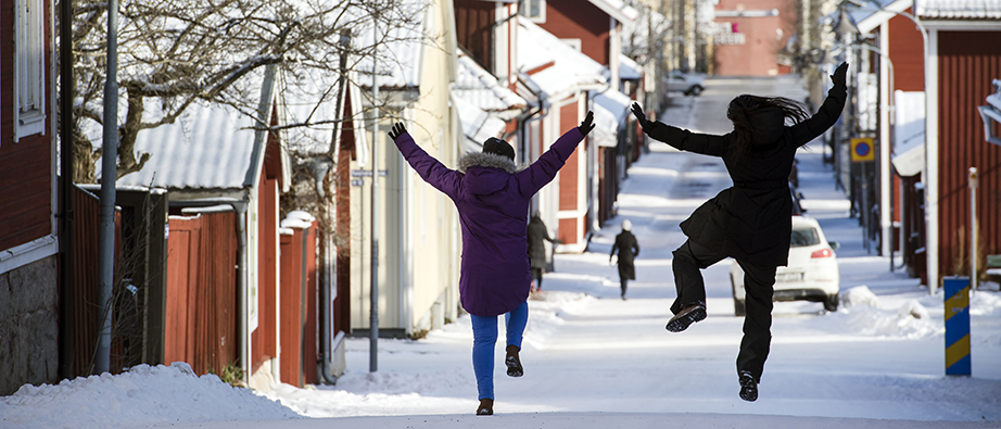 Två glada tjejer hoppar upp. I bakgrunden syns de gamla trästadsdelarna i Falun och det är vinter.